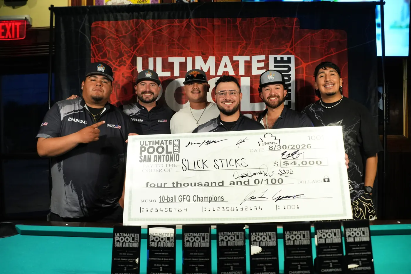 Slick Sticks holding the $4,000 travel-fund check dated 8/30/2026, made out to them as 10-Ball GFQ Champions, in front of the Ultimate Pool League backdrop at Fast Eddie's Braun with the row of trophies in front.