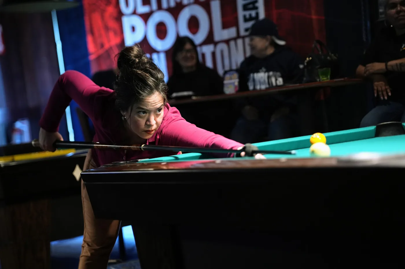 A player in pink stretched across the table for a shot during the 8-Ball qualifier, spectators watching from the tables behind.