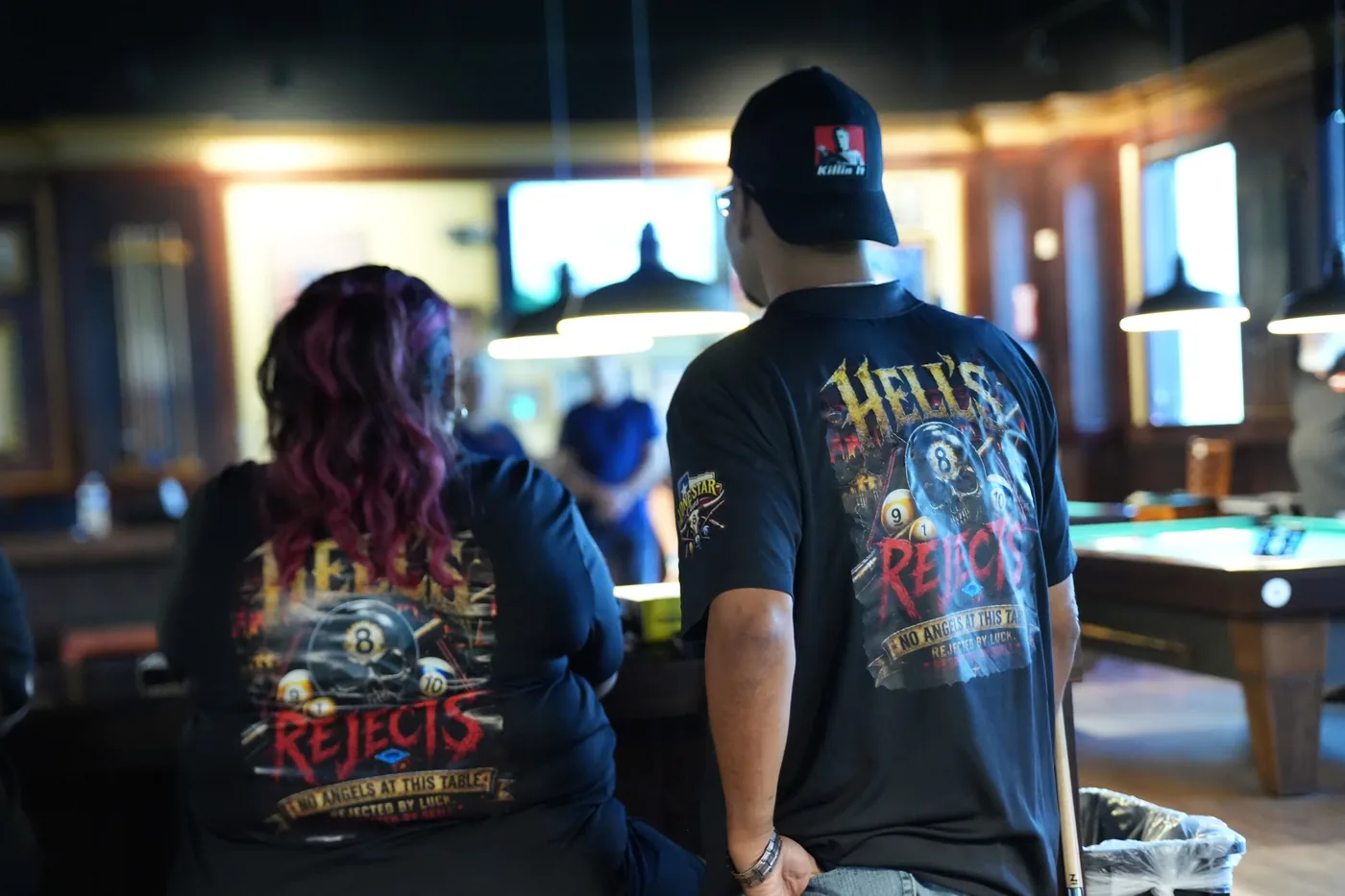 Two Hell's Rejects players seen from behind in their black team jerseys, the full eight-ball design across both backs, watching a match at the 10-Ball qualifier.